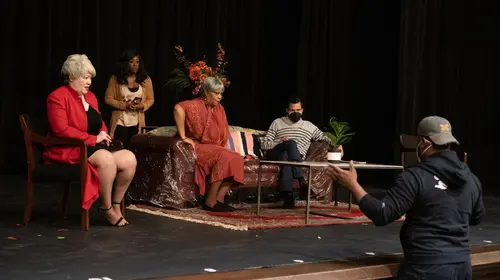 Papa Jennings Is Gone by Aseneth Elizabeth Parker in rehearsal at Obsidian Theatre Festival, March 2021. Two actors seated with crew members beside and behind them. Another crew member stands in front of the stage gesturing with his arms.