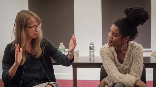 Color photograph of two women in conversation. The woman on the left is white with long, dark blonde hair, wearing glasses and a zippered cardigan sweater over a dark t-shirt. The woman on the right is Black with her many, long braids pulled up on top of her head and wears large round earrings and a loosely woven beige, v-neck sweater.